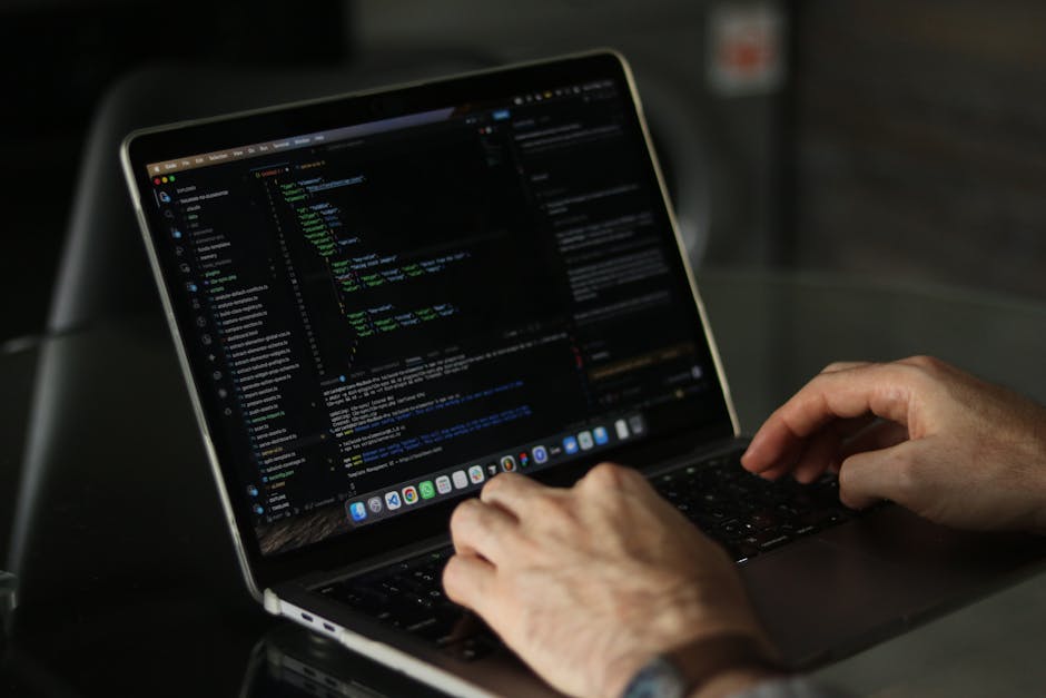 A close-up of a person’s hands operating a laptop on a dark table, with the screen displaying lines of coloured code in a code editor. The individual appears to be engaged in programming or website de