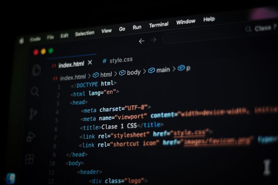 A close-up of a person’s hands operating a laptop on a dark table, with the screen displaying lines of coloured code in a code editor. The individual appears to be engaged in programming or website de