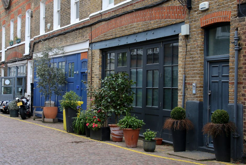 A narrow residential street featuring a row of terraced brick houses with white-framed windows and a small balcony on the upper floor. The foreground showcases a well-maintained garden bed along the b